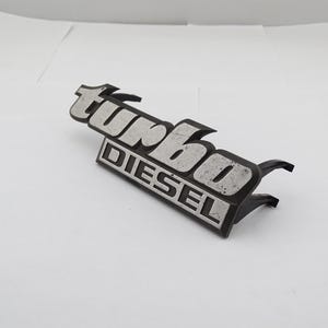 Może przedstawiać: Czarny i srebrny emblemat "Turbo Diesel". Emblemat ma prostokątną podstawę ze słowem "DIESEL" wielkimi literami. Słowo "turbo" znajduje się nad nim, w stylizowanej czcionce.