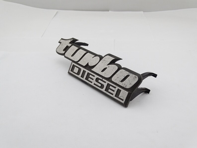 Może przedstawiać: Czarny i srebrny emblemat "Turbo Diesel". Emblemat ma prostokątną podstawę ze słowem "DIESEL" wielkimi literami. Słowo "turbo" znajduje się nad nim, w stylizowanej czcionce.