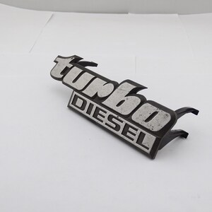 Może przedstawiać: Czarny i srebrny emblemat "turbo DIESEL". Słowo "turbo" jest w stylizowanej czcionce nad słowem "DIESEL". Emblemat ma dwa uchwyty montażowe.