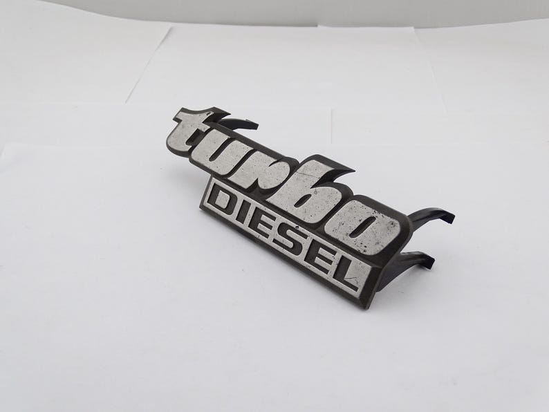 Może przedstawiać: Czarny i srebrny emblemat "turbo DIESEL". Słowo "turbo" jest w stylizowanej czcionce nad słowem "DIESEL". Emblemat ma dwa uchwyty montażowe.