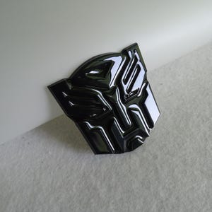 May include: A shiny black Autobot symbol emblem. The badge has a three-dimensional design with reflective surfaces, and is set against a neutral background.