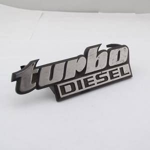 Może przedstawiać: Srebrny i czarny emblemat "turbo DIESEL". Słowo "turbo" jest w stylizowanej czcionce nad słowem "DIESEL". Emblemat jest wykonany z metalu i ma lekko zwietrzały wygląd.