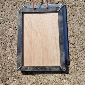 May include: A black metal frame with a wooden insert and a brown leather strap. The frame is hanging on a concrete surface.