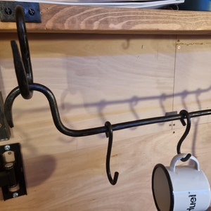 May include: A black metal rack with multiple hooks mounted on a wooden surface. A white mug with black lettering hangs from one of the hooks.