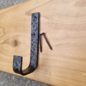 Puede incluir: Un gancho de metal texturizado forjado a mano en forma de J, con dos tornillos negros. El gancho está montado sobre una superficie de madera de color claro. El gancho tiene una apariencia martillada, lo que sugiere un diseño rústico o industrial.
