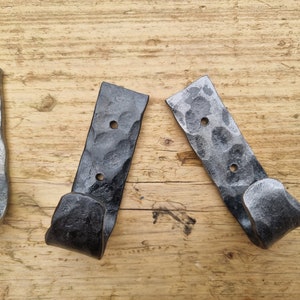 May include: Three black metal hooks with a hammered finish. The hooks are mounted on a wooden surface.
