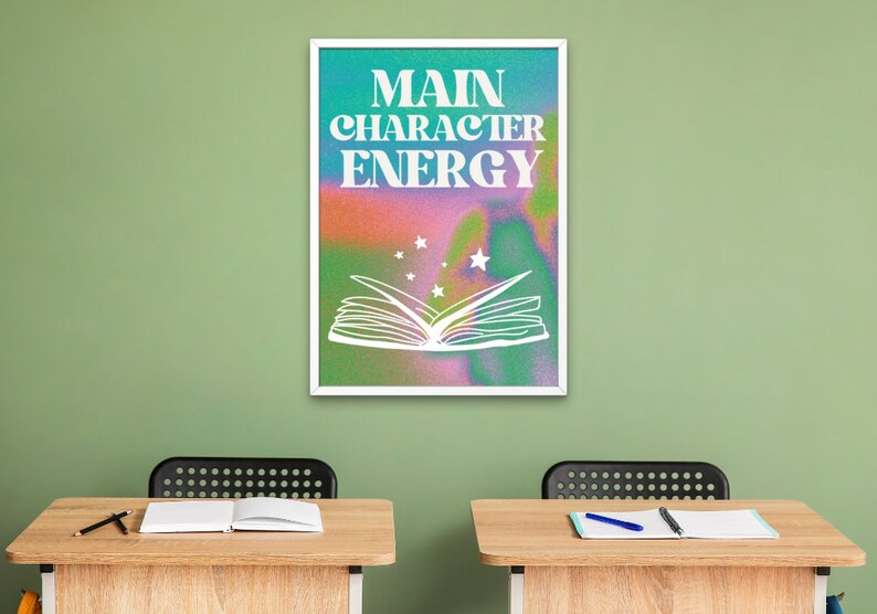 May include: A white framed poster with a colorful background and the text "MAIN CHARACTER ENERGY" in white letters. A white outline of an open book with stars above it is in the center of the poster.