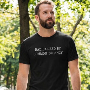 May include: Black crew neck t-shirt with the white text "RADICALIZED BY COMMON DECENCY". The shirt is made of a soft material and is being worn outdoors with a blurred green background.
