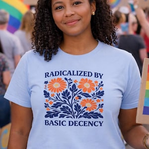 Op de afbeelding: Lichtblauw T-shirt met de tekst "RADICALIZED BY BASIC DECENCY" en een bloemenontwerp in oranje en marineblauw. De persoon houdt een regenboogvlag vast in een menigte.