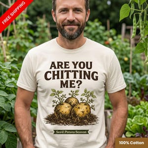 May include: A cream-coloured t-shirt featuring the text "ARE YOU CHITTING ME?" in brown. Below the text is a graphic of three potatoes with sprouts. The words "Seed Potato Season" are below the graphic. The shirt is 100% cotton.