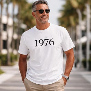 Könnte beinhalten: Ein weißes T-Shirt mit der Jahreszahl "1976" in Schwarz aufgedruckt. Das T-Shirt wird von einer Person getragen, die eine Sonnenbrille und eine Uhr trägt. Die Person steht draußen an einem sonnigen Tag.