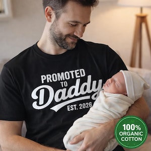 Puede incluir: Camiseta negra con el texto "PROMOTED TO Daddy EST. 2026" en letras blancas. Un bebé recién nacido es sostenido en brazos por una persona que lleva la camiseta. Un círculo verde con el texto "100% ORGANIC COTTON" está en la esquina inferior derecha.