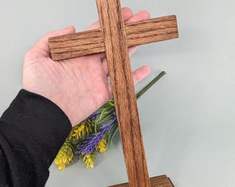 May include: A handcrafted wooden cross with a dark brown finish. The cross is made of rectangular pieces of wood and stands on a square base. The wood grain is visible. The cross is held in a hand with a black sleeve and is next to a small bouquet of flowers.