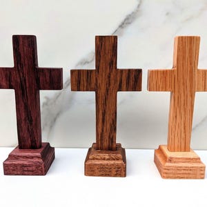 May include: Three wooden crosses in varying shades of brown. The crosses are displayed on individual rectangular bases. The wood grain is visible, and the crosses have a simple, traditional design. The background is a white surface.