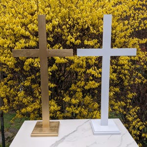 May include: Two wooden crosses, one gold and one white, stand on a white surface against a backdrop of yellow flowers. The gold cross has a square base, while the white cross has a rectangular base. Both crosses are of similar size and design.