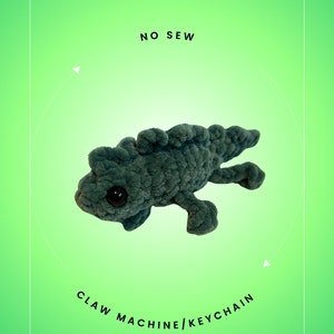 May include: A teal blue crocheted iguana plush toy with large black eyes. The toy is labeled "Iguana Buddy" and "No Sew" and is described as a "Claw Machine/Keychain" created by Brooke Skiver @LilNellieKnits.