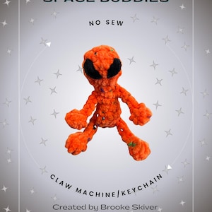 May include: Orange crocheted alien plush toy with black eyes. The toy is labeled "Space Buddies No Sew Claw Machine/Keychain" and "Created by Brooke Skiver @lilnnellieknits".