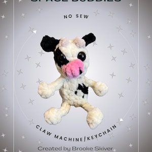 May include: A white and black crocheted cow plush toy with pink ears and nose. The toy is hanging from a string and has a tag that says "Space Buddies No Sew Claw Machine/Keychain Created by Brooke Skiver @lilnellie knits".
