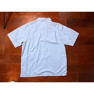 K&ouml;nnte beinhalten: Hellblaues kurz&auml;rmeliges Hemd mit Button-Down-Kragen. Das Hemd ist aus leichtem Stoff gefertigt und hat ein klassisches Design. Das Hemd liegt flach auf einer Holzoberfl&auml;che.