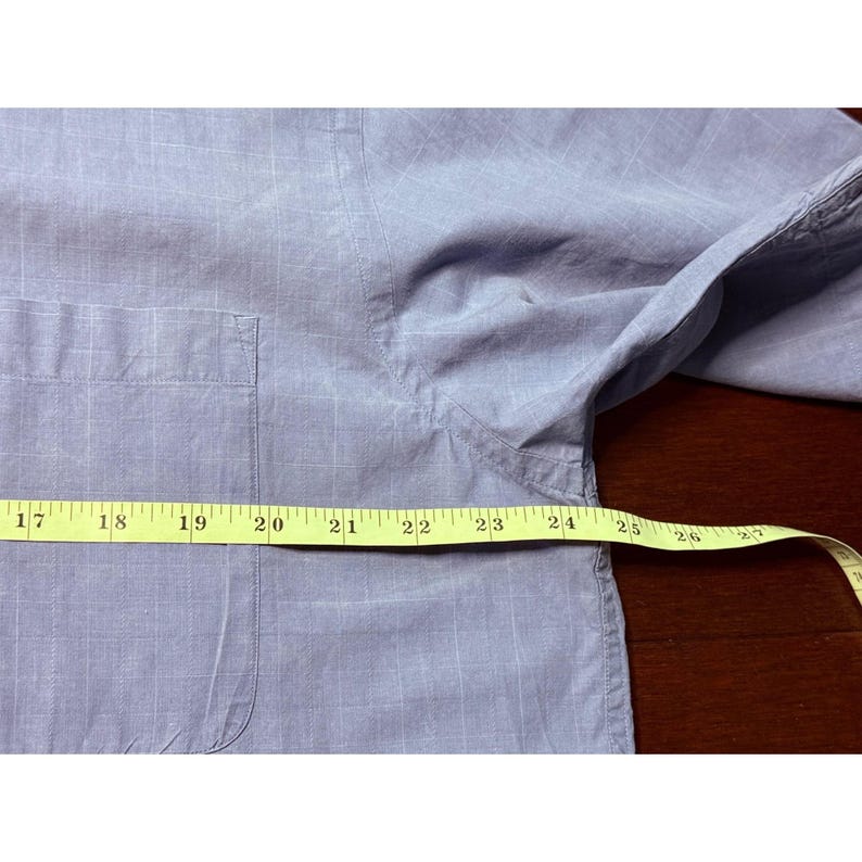 K&ouml;nnte beinhalten: Hellblaues Button-Down-Hemd mit kleiner Brusttasche. Ein gelbes Ma&szlig;band liegt &uuml;ber dem Hemd und zeigt Messungen von 43 bis 66 cm.