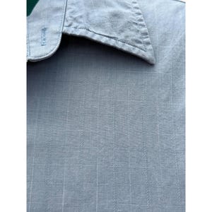 K&ouml;nnte beinhalten: Nahaufnahme eines hellblauen Hemdes mit Knopfleiste und einem Gittermuster. Der Kragen ist sichtbar, und der Stoff hat eine subtile Textur. Das Hemd ist aus leichtem Material. Klassischer Hemdstil.