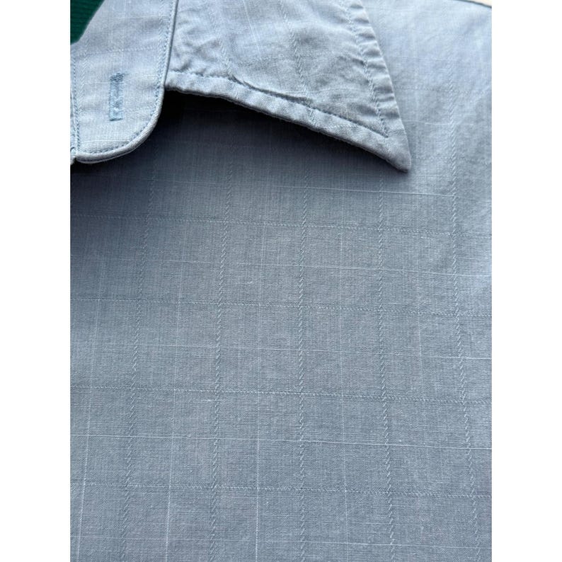 K&ouml;nnte beinhalten: Nahaufnahme eines hellblauen Hemdes mit Knopfleiste und einem Gittermuster. Der Kragen ist sichtbar, und der Stoff hat eine subtile Textur. Das Hemd ist aus leichtem Material. Klassischer Hemdstil.
