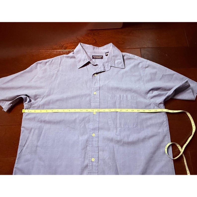 K&ouml;nnte beinhalten: Ein hellblaues Button-Down-Hemd mit Brusttasche und klassischem Kragen. Ein gelbes Ma&szlig;band liegt &uuml;ber dem Hemd. Das Hemd ist aus leichtem Stoff gefertigt.