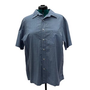 K&ouml;nnte beinhalten: Hellblaues kurz&auml;rmeliges Button-Down-Hemd mit klassischem Kragen und einer einzelnen Brusttasche. Das Hemd ist aus leichtem Stoff gefertigt und hat wei&szlig;e Kn&ouml;pfe auf der Vorderseite. Das Hemd wird auf einer Schaufensterpuppe pr&auml;sentiert.