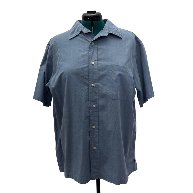K&ouml;nnte beinhalten: Hellblaues kurz&auml;rmeliges Button-Down-Hemd mit klassischem Kragen und einer einzelnen Brusttasche. Das Hemd ist aus leichtem Stoff gefertigt und hat wei&szlig;e Kn&ouml;pfe auf der Vorderseite. Das Hemd wird auf einer Schaufensterpuppe pr&auml;sentiert.