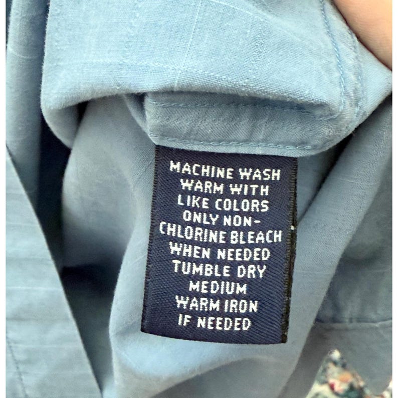 K&ouml;nnte beinhalten: Nahaufnahme eines hellblauen Hemdes mit einem Pflegeetikett. Das Etikett, wei&szlig;er Text auf marineblauem Hintergrund, lautet: "MASCHINENW&Auml;SCHE WARM MIT &Auml;HNLICHEN FARBEN NUR NICHT-CHLORBLEICHE WENN N&Ouml;TIG TROCKNEN IM TROCKNER MITTLERE W&Auml;RME B&Uuml;GELN WARM WENN N&Ouml;TIG."