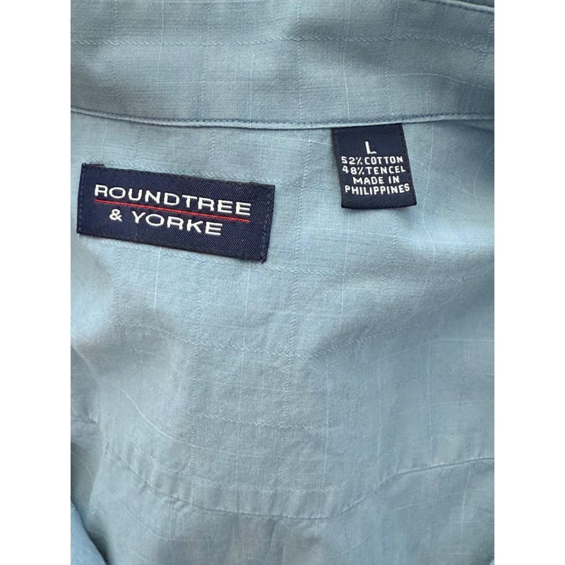 K&ouml;nnte beinhalten: Nahaufnahme eines hellblauen Hemdes mit dezentem Karomuster. Das Hemd hat zwei marineblaue Etiketten. Auf einem Etikett steht "ROUNDTREE & YORKE" in wei&szlig;er Schrift. Das andere Etikett gibt die Gr&ouml;&szlig;e L und die Herstellung in den Philippinen an.