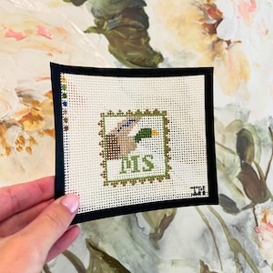 May include: A small, square needlepoint featuring a duck design within a green and brown border. The word "DMS" is stitched in green. The needlepoint is set against a black border.
