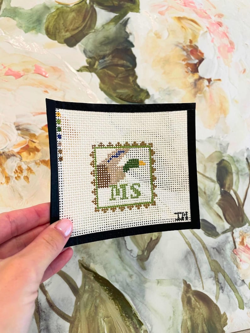 May include: A small, square needlepoint featuring a duck design within a green and brown border. The word "DMS" is stitched in green. The needlepoint is set against a black border.