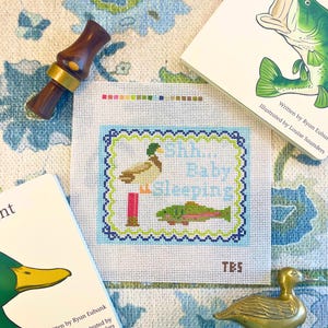 May include: A needlepoint canvas with a duck and fish design, the text "Shh... Baby Sleeping", and the initials TBS. The image also includes two children's books, a duck call, and a brass duck figurine. The canvas is on a blue floral patterned background.