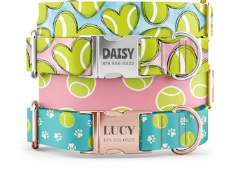 May include: Three dog collars with different patterns. The top collar is light blue with white hearts and green tennis balls. The middle collar is pink with green tennis balls. The bottom collar is teal with white paw prints and green tennis balls. Each collar has a metal buckle with a name and phone number engraved on it. The top buckle says "DAISY 419.555.0322", the middle buckle says "DAISY 419.555.0322", and the bottom buckle says "LUCY 419.555.0322".