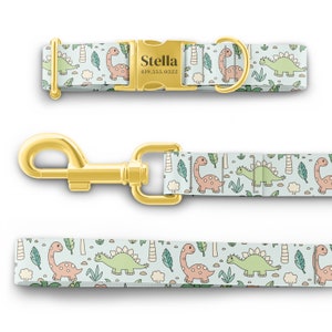 May include: A light blue dog collar and leash with a white background and a pattern of green and pink dinosaurs. The collar has a gold buckle with the name "Stella" and a phone number engraved on it.