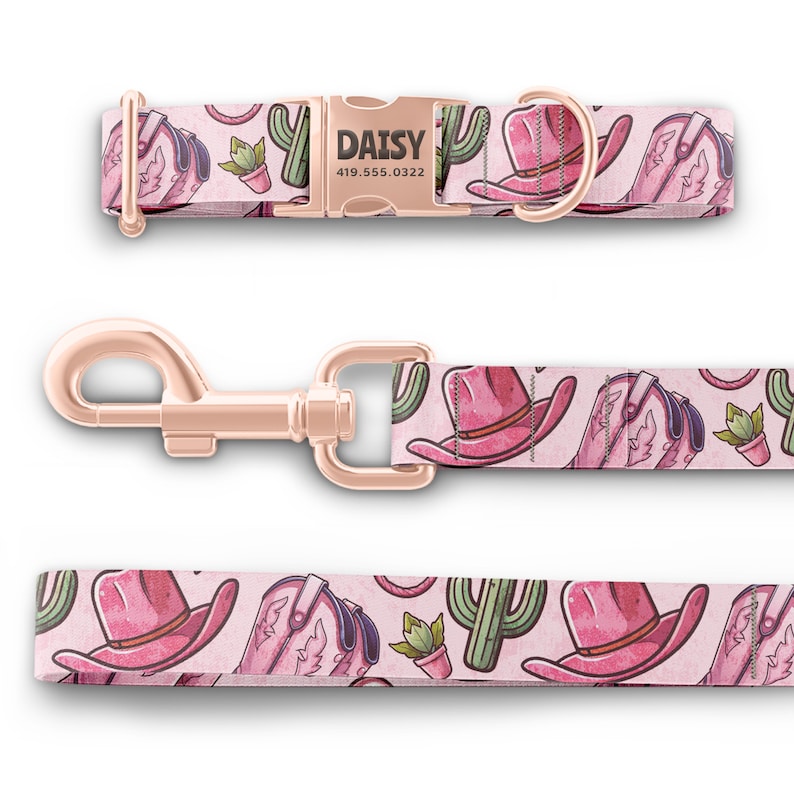 May include: A pink dog collar and leash with a western theme. The collar has a rose gold buckle with the name "DAISY" and a phone number. The leash and collar feature a repeating pattern of pink cowboy hats, boots, and cacti.