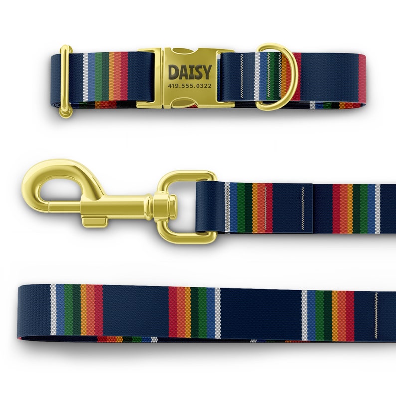 May include: A navy blue dog collar and leash set with a rainbow stripe design. The collar has a gold-colored buckle with the name "DAISY" and a phone number. The leash has a gold-colored clasp.