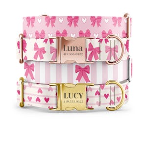 May include: Three pink and white dog collars with gold and rose gold buckles. The collars feature different patterns: pink bows on a white background, pink and white stripes, and pink hearts on a white background. The buckles are engraved with the names "Luna" and "Lucy" and phone numbers.