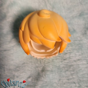 May include: A yellow plastic doll hair piece with a center part and a side part. The hair is styled in a short, choppy bob.