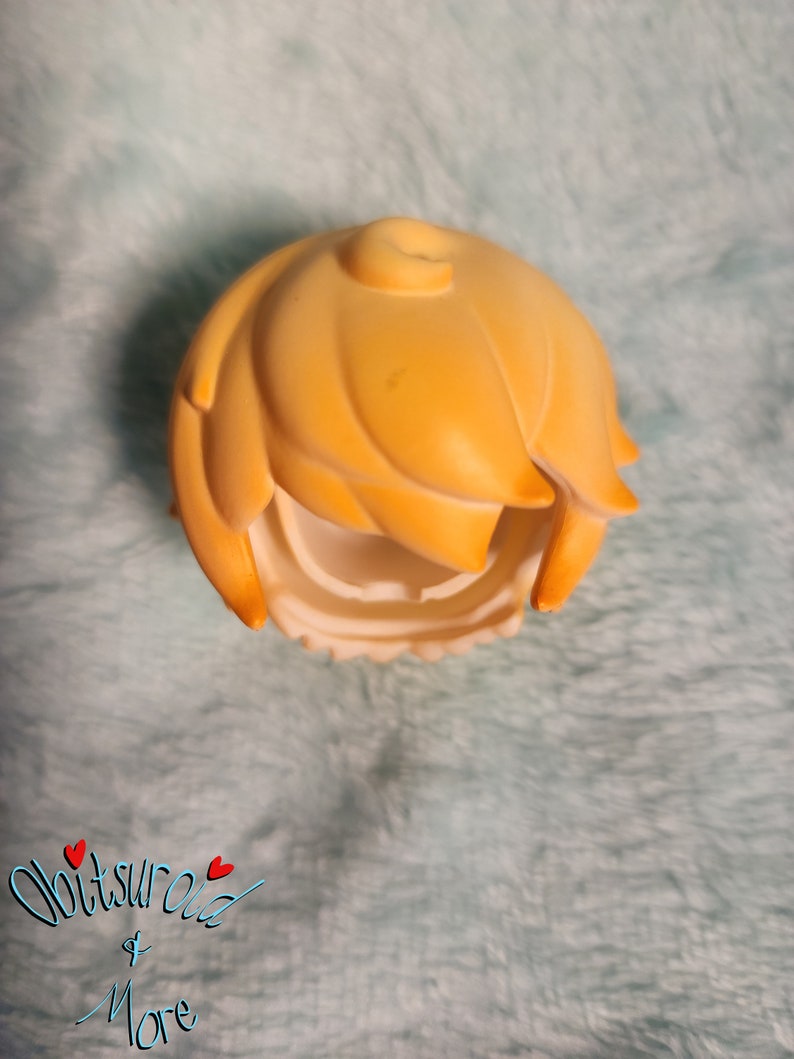 May include: A yellow plastic doll hair piece with a center part and a side part. The hair is styled in a short, choppy bob.