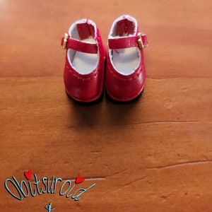 May include: A pair of red leather doll shoes with gold buckles. The shoes have a white lining and are on a wooden surface.