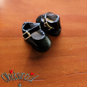 May include: A pair of black doll shoes with a buckle closure. The shoes are made of a textured material and have a rounded toe.
