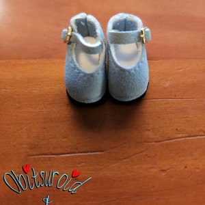May include: A pair of light blue doll shoes with gold buckles. The shoes are made of a soft, suede-like material and have a white lining.