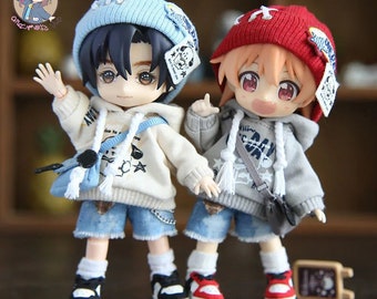 May include: Two small dolls wearing grey hooded sweatshirts with white trim, blue denim shorts, and sneakers. One doll is wearing a blue beanie with a white star and a white dog graphic. The other doll is wearing a red beanie with a white star and a white dog graphic.