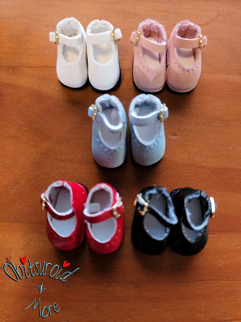 May include: Six pairs of miniature doll shoes in various colors, including white, pink, blue, red, and black. The shoes have a simple design with a buckle strap and a rounded toe.