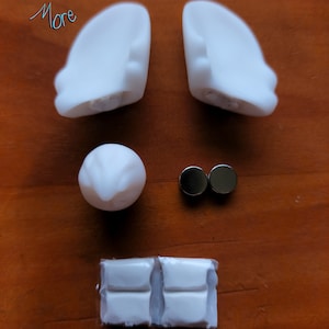 May include: A set of white plastic doll ears, a white plastic nose, two silver magnets, and two white plastic pieces with a textured surface.