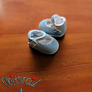 May include: A pair of light blue suede doll shoes with gold buckles. The shoes are on a wooden surface.