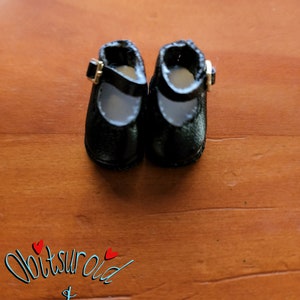 May include: A pair of black leather doll shoes with a buckle closure. The shoes are on a wooden surface.