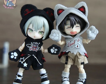 May include: Two small dolls wearing hooded outfits with paw print details. The doll on the left is wearing a black hooded outfit with a red and black plaid trim and a bear graphic on the front. The doll on the right is wearing a gray hooded outfit with a red and black plaid trim and a graphic that says "Good at".